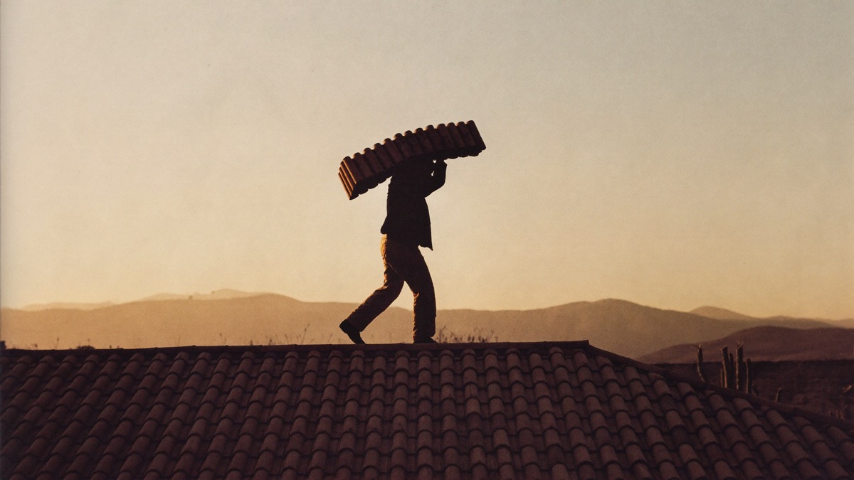 Roofer carrying barrel tile along a ridge line at sunrise, silhouetted against desert mountains