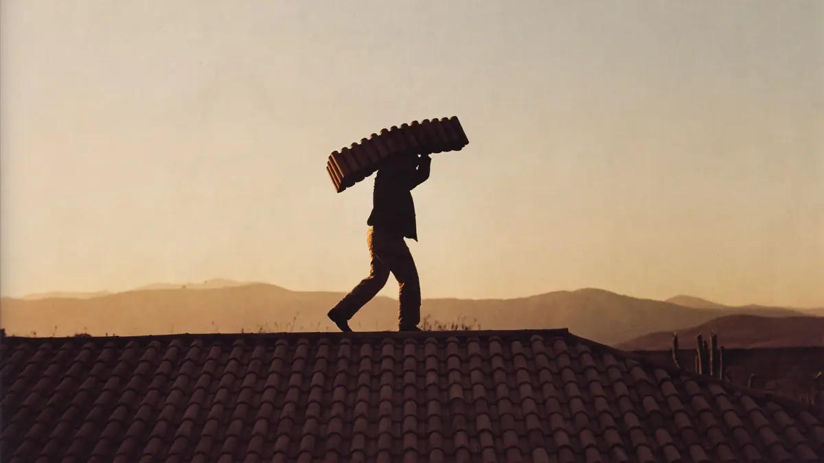 Roofer carrying barrel tile along a ridge line at sunrise, silhouetted against desert mountains